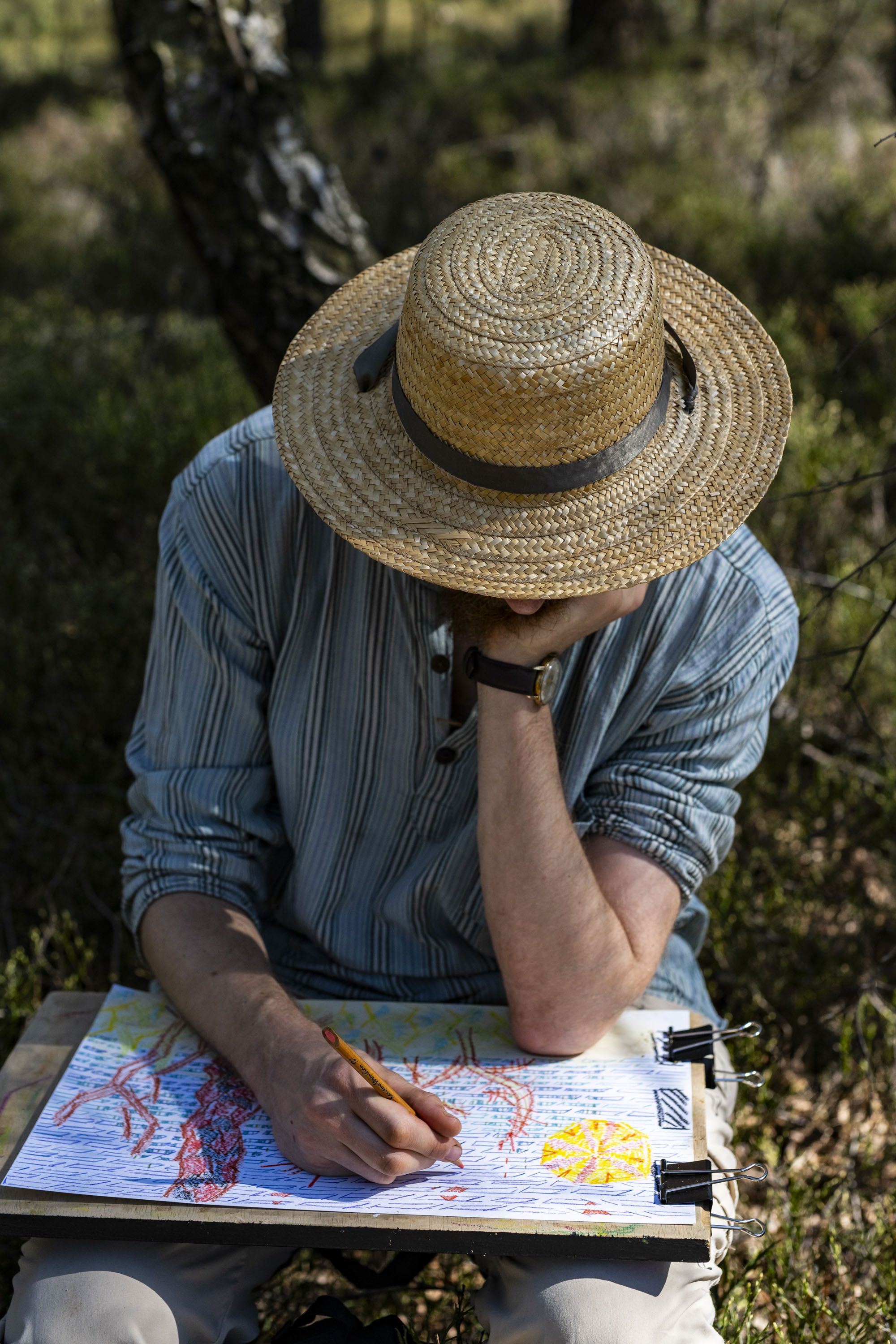 A man wearing a straw hat is focused on drawing on a piece of paper, surrounded by natural scenery.